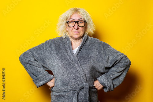 Smiling old woman in gray bathrobe holds hands on belt on yellow background.