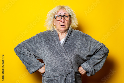 Surprised old woman in gray bathrobe holds hands on belt on yellow background.
