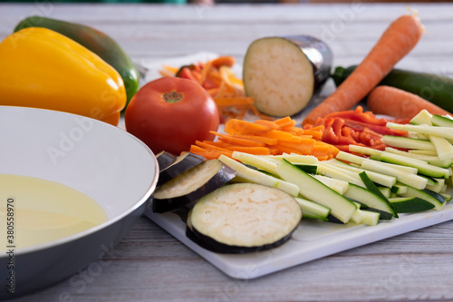 White pan with olive oil on wooden table with a mix of sliced vegetables, carrots, zucchini, eggplant and peppers ready to be cooked - healthy eating vegetarian vegan concept photo