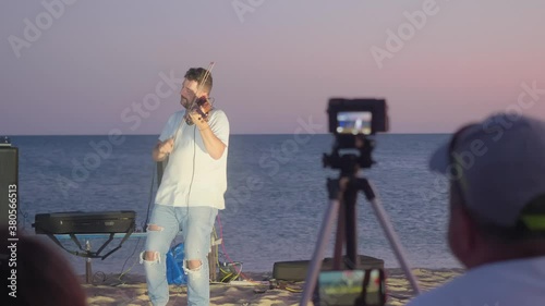 Man plays the violin standing near the sea. Camera man makes the video and listens. Amazing summer sunset.