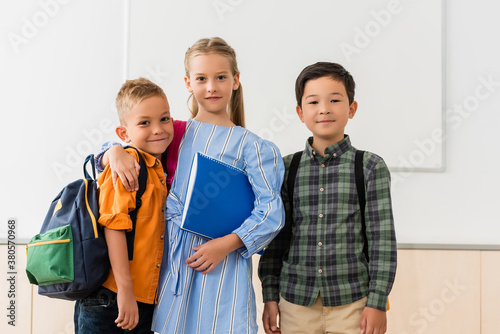 Schoolgirl with notebook embracing classmate near asian friend in classroom