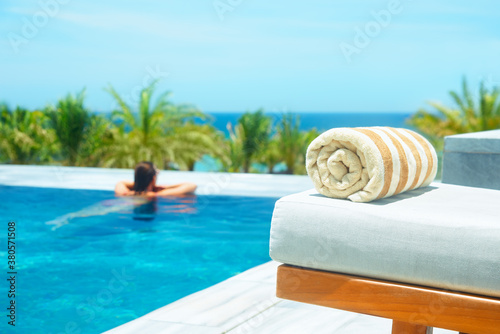 Fototapeta Naklejka Na Ścianę i Meble -  Rolled up towel on lounge chair by luxury outdoor pool to background of palm trees and ocean. Focus on foreground