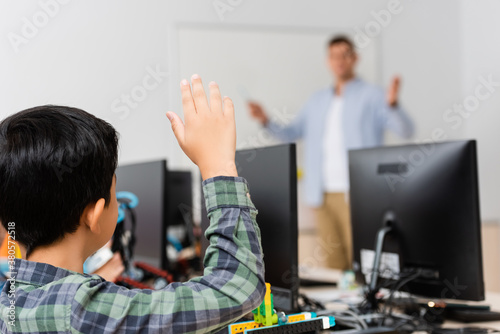 Selective focus of schoolboy with raised hand sitting near robot and computers during lesson in stem school