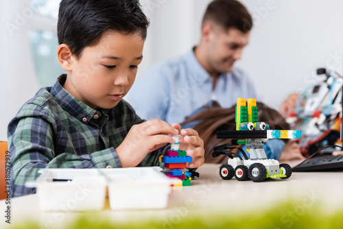 Selective focus of asian schoolboy with building blocks modeling robot near friend and teacher in school