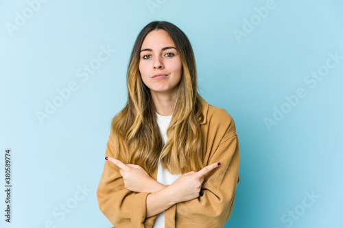 Young caucasian woman isolated on blue background points sideways, is trying to choose between two options. © Asier