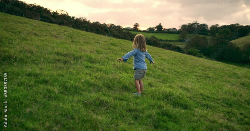 A preschooler is running and spinning in a field at sunset