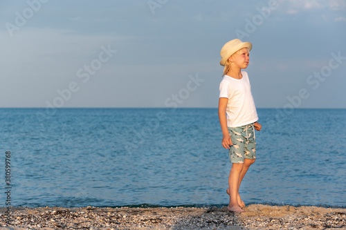 Boy in straw hat walks along the seashore in rays of sunset. Carelessness