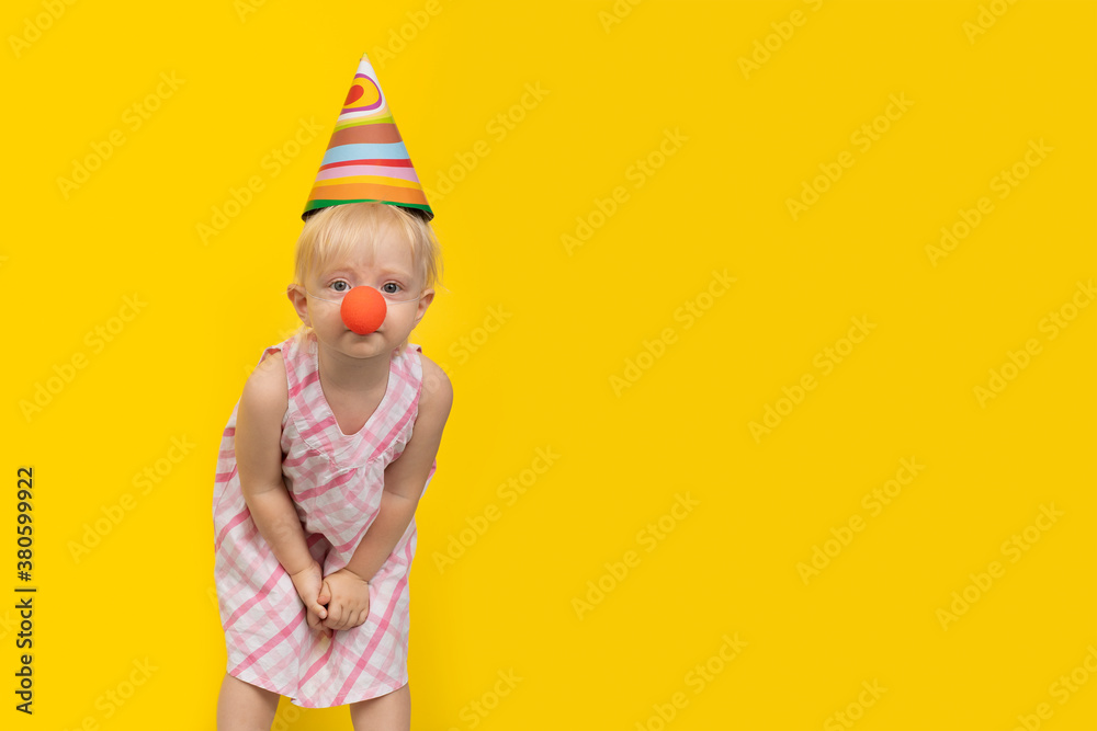 Cute fair-haired girl in festive hat and clown nose looking at camera. Holiday or birthday party concept. Copy space.