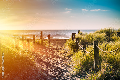 Golden sunset over sandy pathway with grass reeds and wooden posts on each side leading to a beautiful sea bay