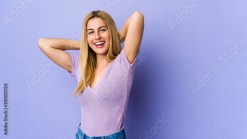 Young blonde woman isolated on purple background feeling confident, with hands behind the head.