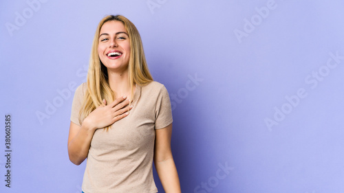 Young blonde woman isolated on purple background laughs out loudly keeping hand on chest.