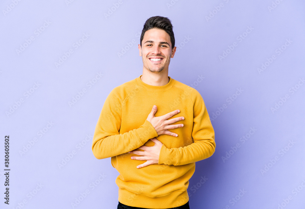 Young caucasian man isolated on purple background laughs happily and has fun keeping hands on stomach.