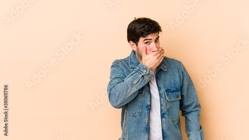 Young man isolated on beige background covering mouth with hands looking worried. © Asier