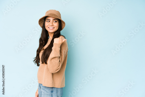 Young indian woman wearing a hat isolated on blue background points with thumb finger away, laughing and carefree.