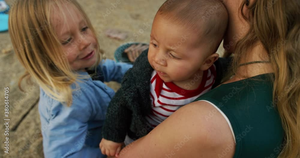 A young mother is sitting on the beach with her preschooler embracing his baby sibling