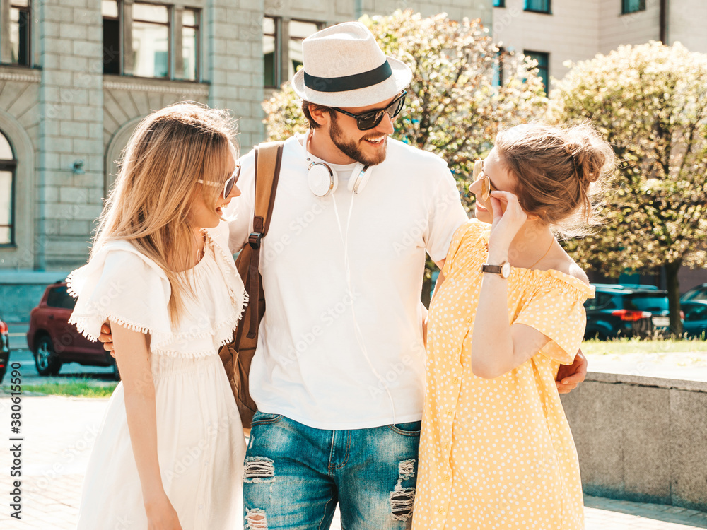 Group of young three stylish friends posing in the street. Fashion man and two cute female dressed in casual summer clothes. Smiling models having fun in sunglasses.Cheerful women and guy chatting
