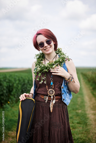 Young hippie woman with short red hair, wearing boho style clothes, sunglasses, with flower wreath on neck, standing on green field, holding guitar. Indie musician in countryside. Eco tourism concept.