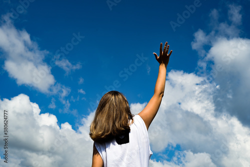 A woman stands with her back to the photographer in a white T-shirt and against a blue sky with clouds. Reaches her hand to the sky. 