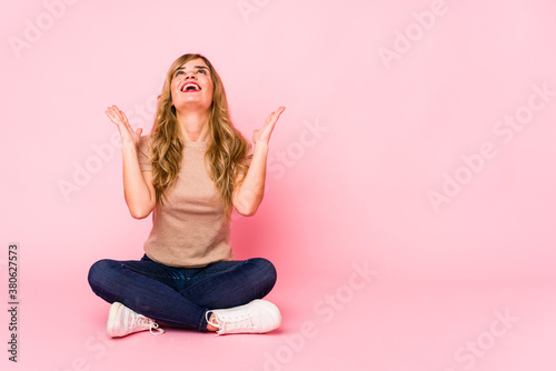 Young blonde caucasian woman sitting on a pink studio screaming to the sky, looking up, frustrated. © Asier