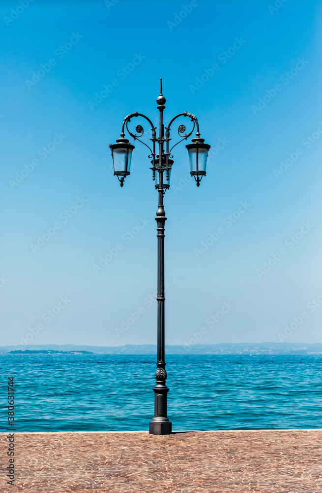 Lantern with three bulbs on the stone sidewalk near the lake water. The water and the sky are blue. Mountains can be seen on the opposite shore of the lake