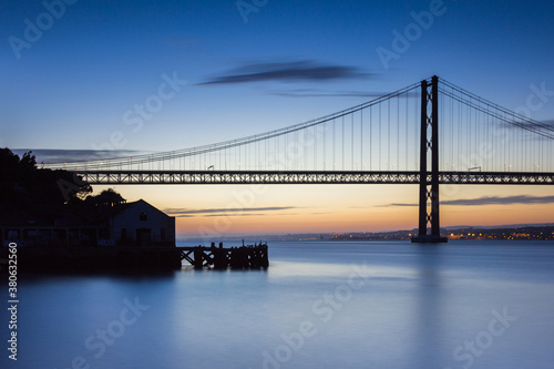 Long exposure photo with blue and silk water and silhouette of the 25 April Bridge in Lisbon, Portugal on a blue hour after sunset.