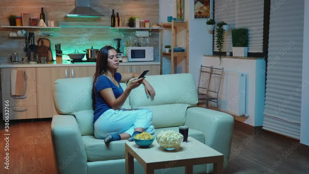 Young lady in pijamas watching Tv in living room sitting on sofa. Bored, home alone late at night woman relaxing watching tv lying on comfortable couch holding remote control changing TV channels. Young lady in pijamas watching Tv in living room sitting on sofa. Bored, home alone late at night woman relaxing watching tv lying on comfortable couch holding remote control changing TV channels.
