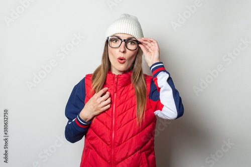 Young woman in a red vest, a hat and glasses with a surprised face on a light background