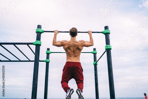 Back view of muscular male athlete training biceps while doing pull ups on sports playground, good looking man with perfect body spending morning time for doing workout exercising on bars