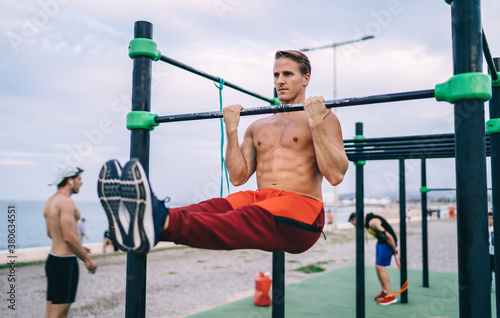 Powerful caucasian male athlete training outdoors keeping muscular strength and vitality on free time, sportsman having street workout on horizontal bars slimming and reaching goals losing weight