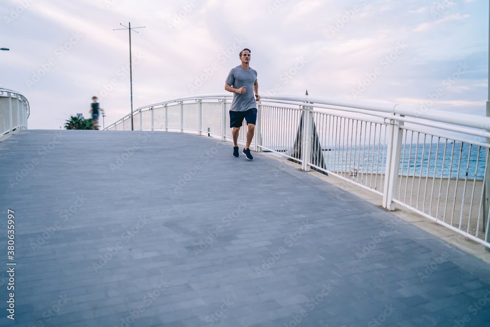 Serious strong male runner in activewear training cardio outdoors in morning slimming and getting energy, handsome caucasian sportsman jogging on bridge keeping healthy lifestyle for slimming