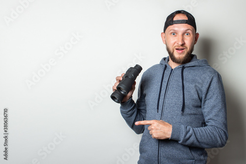 Young man with a surprised face in a hoodie and a cap holds binoculars in his hands on a light background