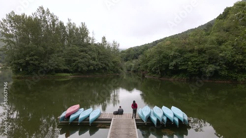 Woman Walking Towards A Man Sitting At The Edge Of The Jetty In Lake Surrounded By Lush Green Trees In Valdemurio Reservoir, Las Agueras, Asturias, Spain - drone shot photo