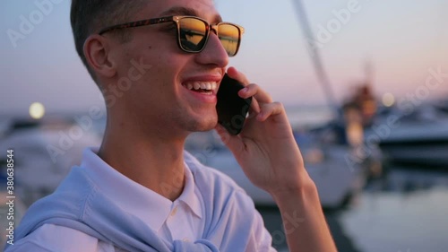 blond guy in sunglasses reflecting orange sunset talks on modern smartphone standing on seafront slow motion closeup