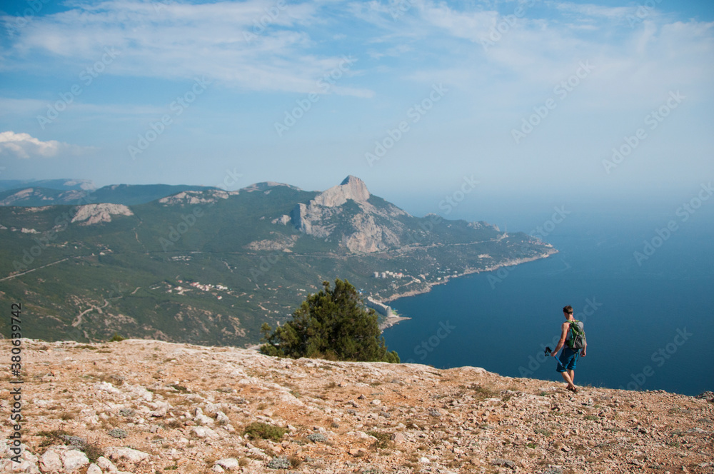 A man with a backpack and trekking poles walking on the edge of a cliff above the sea. Hiking in the mountains high above the sea.
