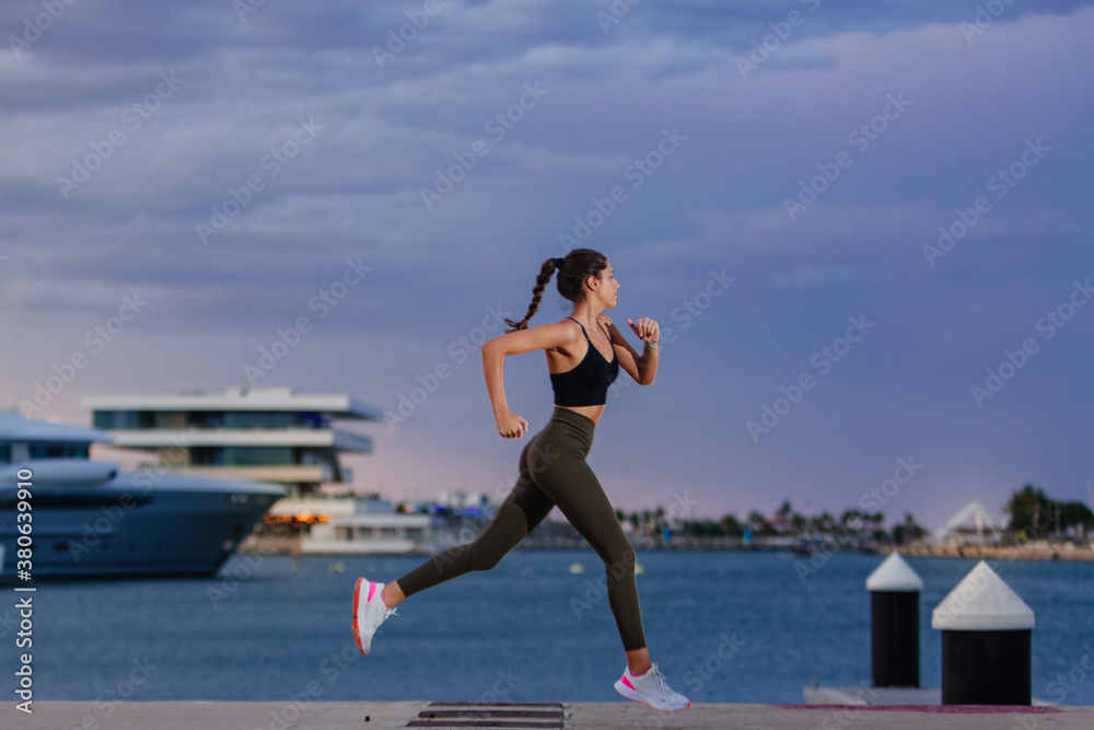 Runner dressed in sportswear runs along the sea in a harbor with docked boats. The girl is young and has an athletic body