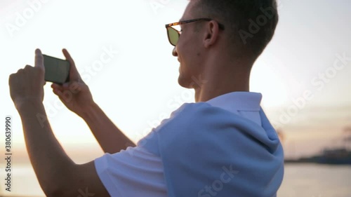 delighted guy in white t-shirt and black sunglasses makes photos of summer sunset walking and smiling slow motion