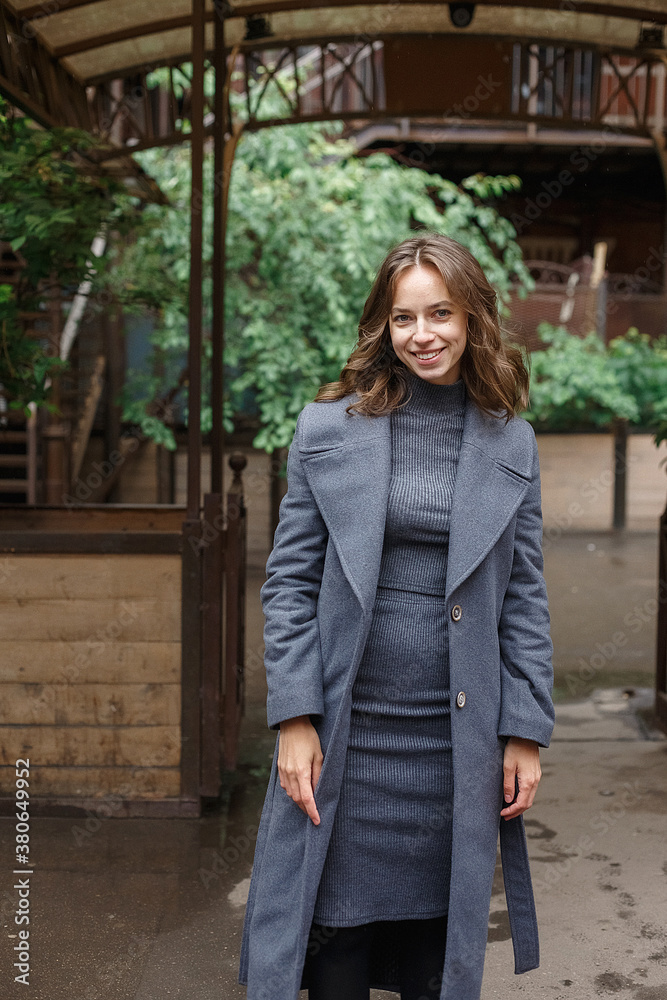 Fototapeta premium young happy smiling pretty girl in gray coat and turtleneck poses outdoors. Street style portrait photo session of attractive female, urban photoshoot of beautiful elegant model