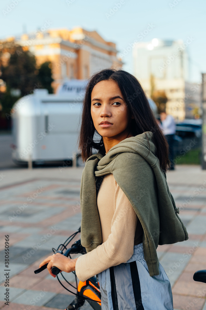 Fototapeta premium Street portrait of a stylish latin woman in casual clothes with a bicycle standing on the street and looking away. Vertical.