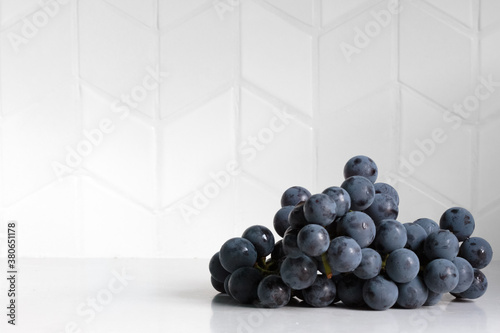 macro shot with natural lighting of a bunch of concord grapes with water droplets on a white stone countertop and a chevron pattern tile backsplash with shadow and copy space