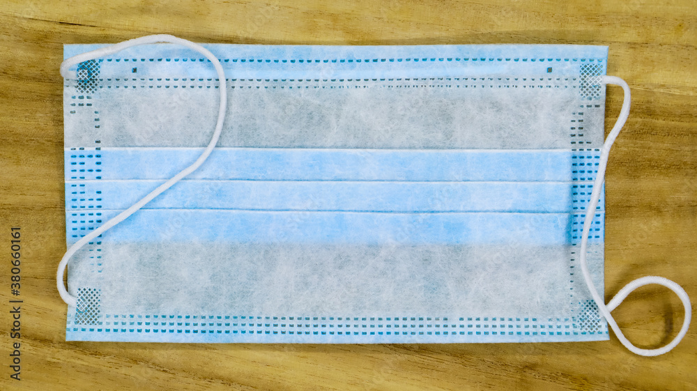 One 3-ply disposable surgical face mask with rubber pads covering the mouth and nose on a wooden table. The concept of protection from bacteria, healthcare and medicine. COVID-19. Dangerous virus. One 3-ply disposable surgical face mask with rubber pads covering the mouth and nose on a wooden table. The concept of protection from bacteria, healthcare and medicine. COVID-19. Dangerous virus.