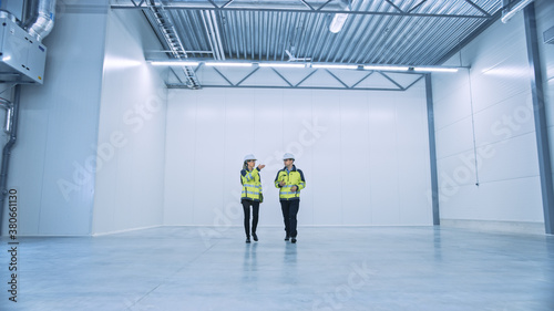 Two Engineers Talking, Walking Across Empty Factory Floor, inspecting, Planning Where All the Machinery Would go. Professionals Industrial Designers Strategizing about New Manufacturing Plant Design © Gorodenkoff