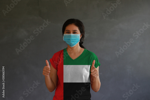 Woman wearing hygienic mask and wearing UAE flag colored shirt with thumbs up with both hands on dark wall background. photo