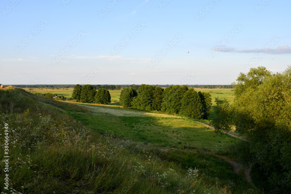 Obraz premium A view from the top of a grassy hill showing vast fields, meadows, and pasturelands, a path of small forest or moor, as well as a cloudless summer sky with a single ballon flying over it in Poland