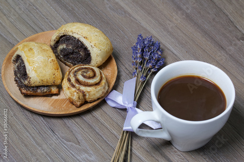 Baked buns stuffed with poppy and cinnamon. Lie on a plate. Next to it is a cup of coffee and a bouquet of lavender.