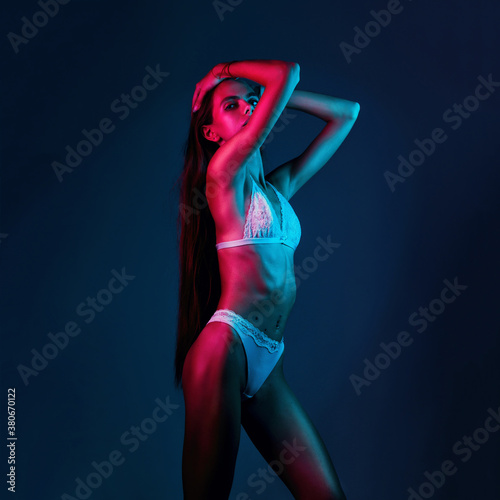 Skinny fashion model wear beige erotic lingerie in colorful bright neon uv red and blue lights, posing in studio hold long hair copy space