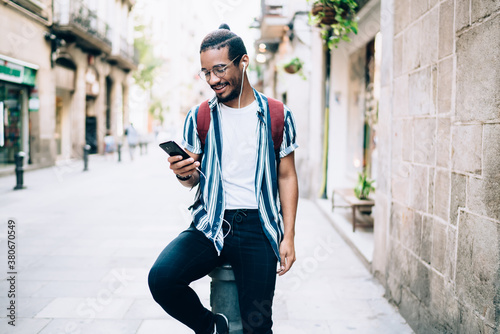 Smiling african american trendy dressed hipster guy in eyewear recreating outdoors using smartphone and sound application for enjoy music, positive dark skinned male listening audiobook on street