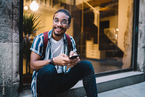 Bearded ethnic man browsing smartphone on stair and looking at camera