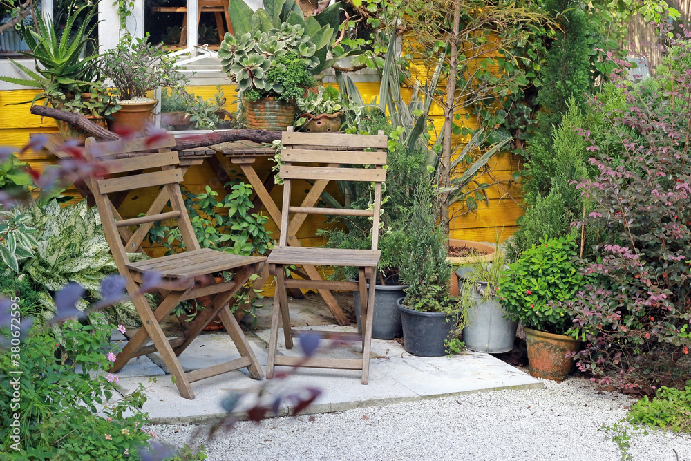 A pair of wooden chairs in the alcove patio of a beautiful tropical garden of flowers and pot plants with a yellow building wall in the background