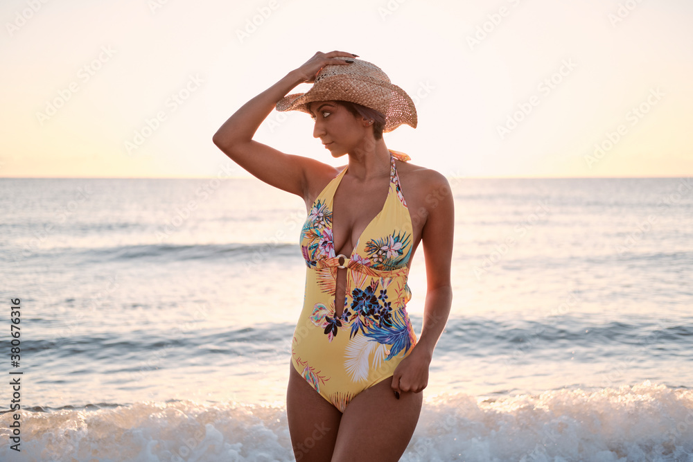 Latin girl standing near of the shore of a deserted beach touching her hat. Wearing a yellow swimsuit and a hat. Latin girl standing near of the shore of a deserted beach touching her hat. Wearing a yellow swimsuit and a hat.