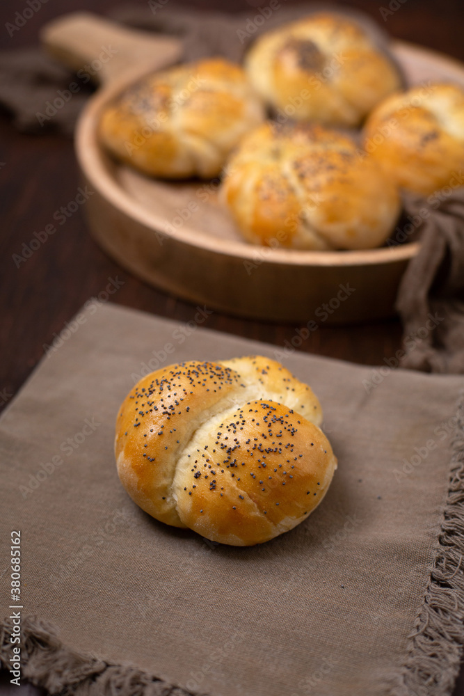 Homemade Kaiser Roll Topped with Poppy Seeds on a Brown Napkin; More in a Round Wooden Bowl in the Background on a Wooden Table Homemade Kaiser Roll Topped with Poppy Seeds on a Brown Napkin; More in a Round Wooden Bowl in the Background on a Wooden Table